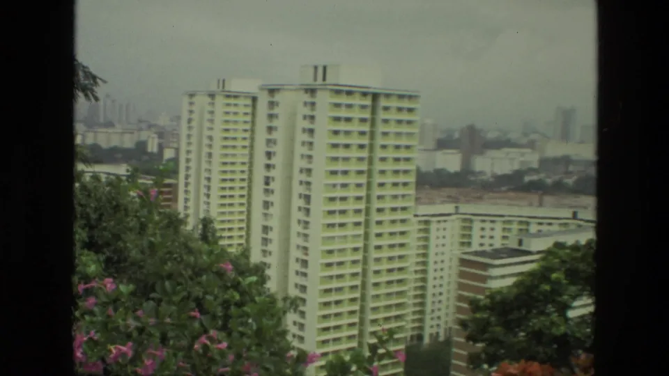 1974 8mm view in India of a tall off-white apartment tower, lower residential blocks, trees, and a hazy distant skyline.