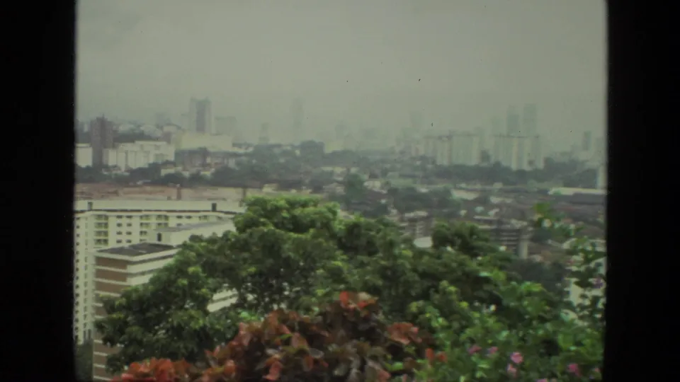1974 India 8mm frame looking over trees and residential blocks toward a hazy urban skyline with distant high-rises.