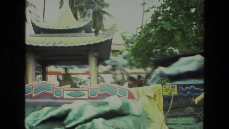 1974 India 8mm frame showing a colorful garden display with a pavilion roof, painted railings, sculpted forms, foliage, and background visitors.