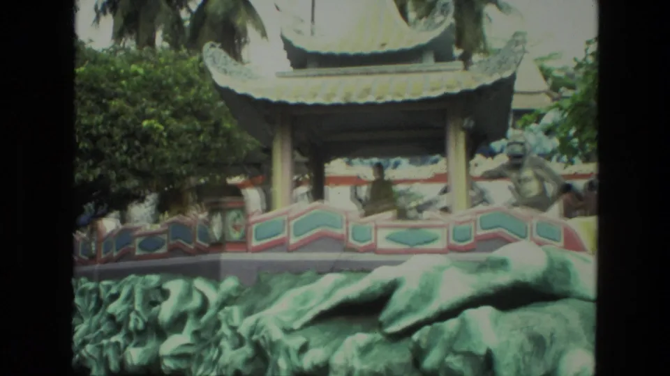 1974 India 8mm frame of a colorful garden tableau with sculpted forms, decorative railing, pavilion rooflines, trees, and a background visitor.