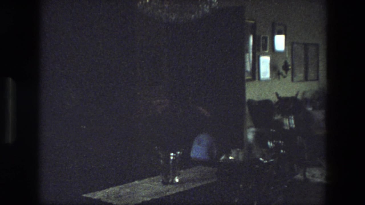 Dim living room with framed pictures, chairs, a table, and a seated person in shadow.
