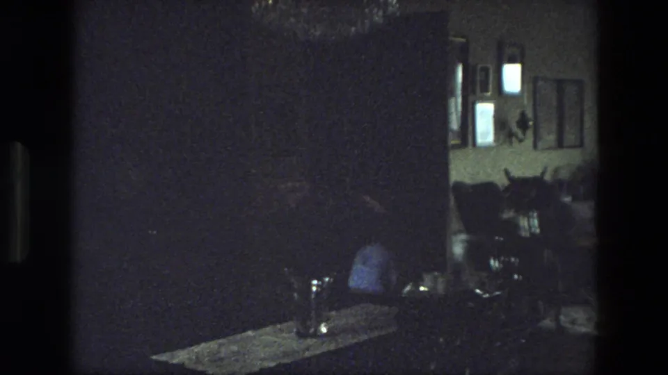 1974 India home-movie frame of a dark living room with wall pictures, furniture, and a seated person partly visible in low light.