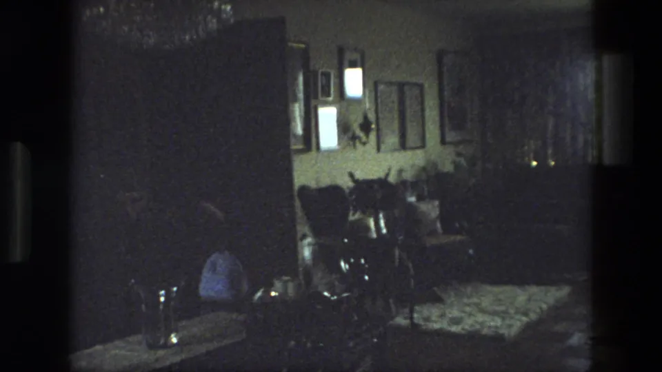 1974 India home interior with framed pictures, chairs, a patterned floor covering, and a seated figure partly obscured by shadow.