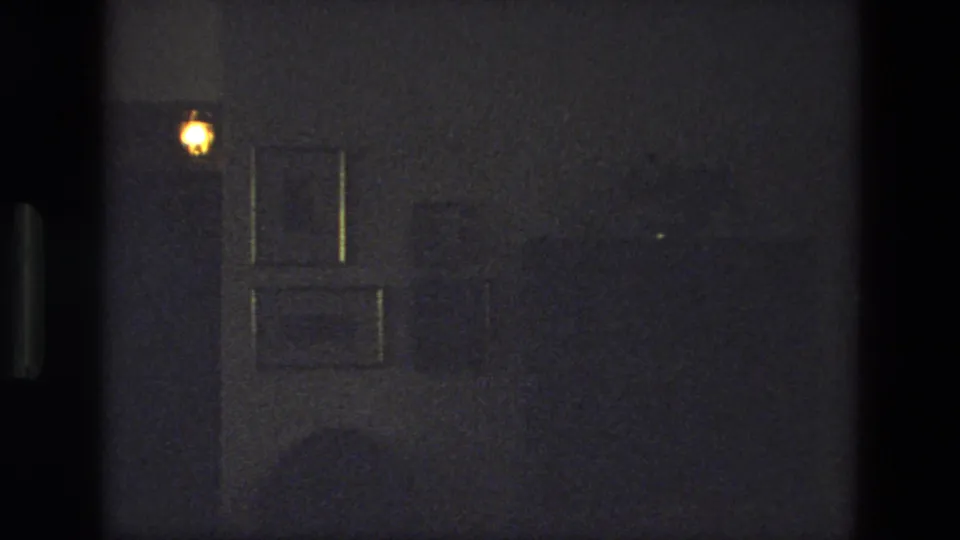 Dark 1974 India interior frame with a small warm light and a paneled door or wall surface barely visible.