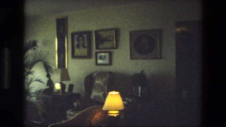 1974 India 8mm frame showing a private living room with portraits, wall pictures, a plant, upholstered furniture, and lamp light.