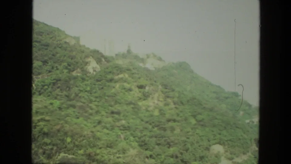 1974 India 8mm view of a lush hillside ridge with rocky breaks and faint distant structures at the crest.
