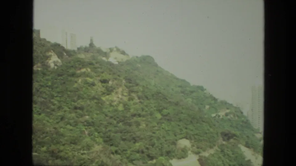 1974 India 8mm frame showing a wooded mountain slope with distant high-rise buildings partly visible through haze.
