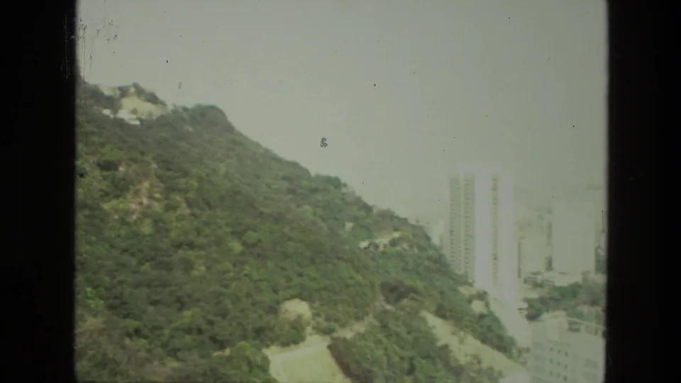 1974 India 8mm view of a lush hill, pale rocky outcrops, and distant high-rise buildings beyond the slope.