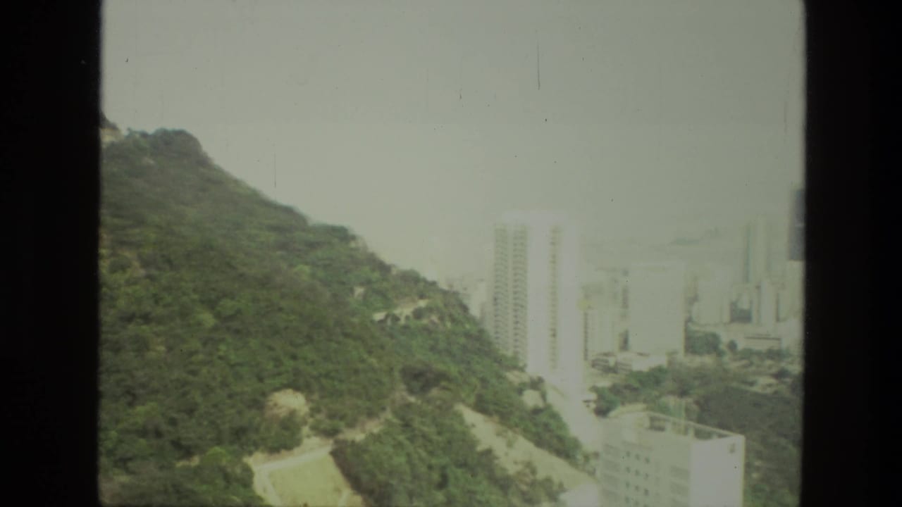 1974 India home-movie frame showing a green hillside, winding road, and distant high-rise buildings through haze.