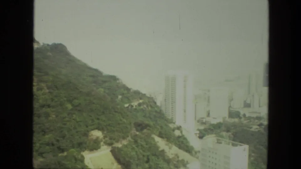 1974 India home-movie frame showing a green hillside, winding road, and distant high-rise buildings through haze.