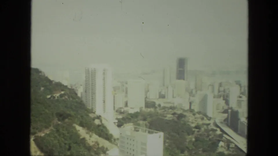 Elevated 1974 India city view with green hillside at left, clustered buildings, haze, and visible 8mm frame edge.