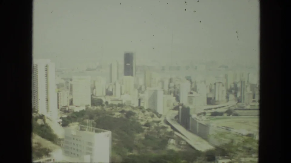 1974 India frame showing a hazy skyline, tower blocks, curving road, and greenery from an elevated home-movie viewpoint.
