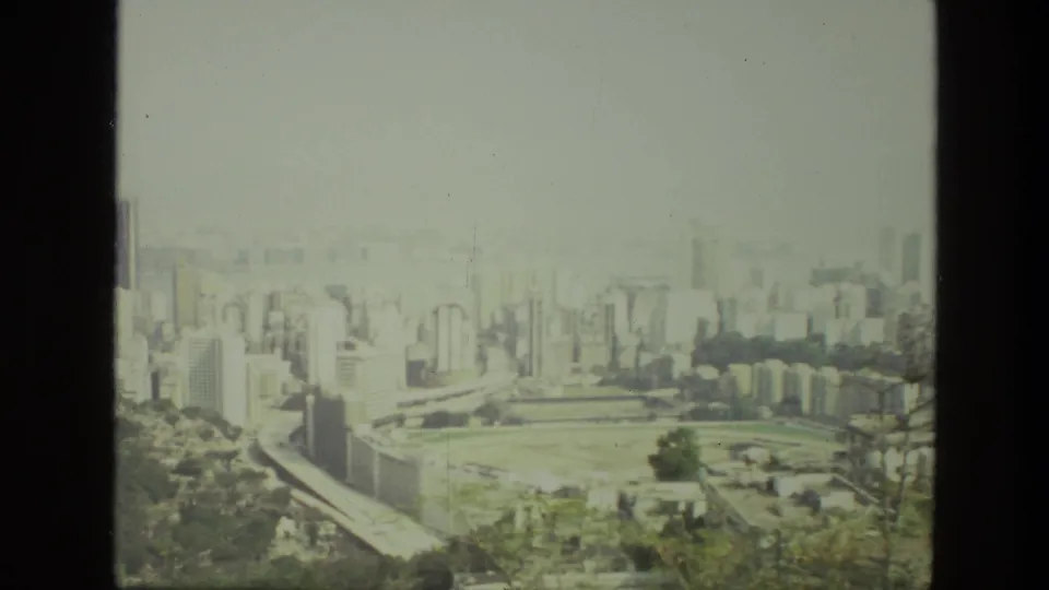 1974 India 8mm frame showing a hazy urban skyline, dense buildings, green open ground, and trees from a high vantage point.