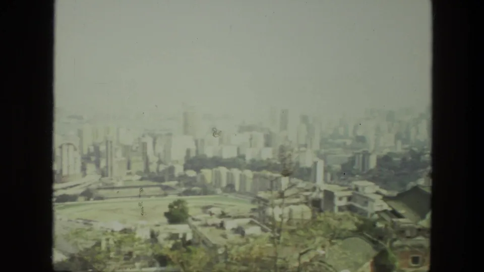 1974 India home-movie frame with a broad city panorama, pale high-rises, clustered buildings, and a large open field or stadium area.