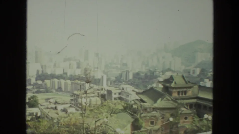 1974 India 8mm frame where tiled-roof architecture and hillside trees sit before a hazy modern skyline.