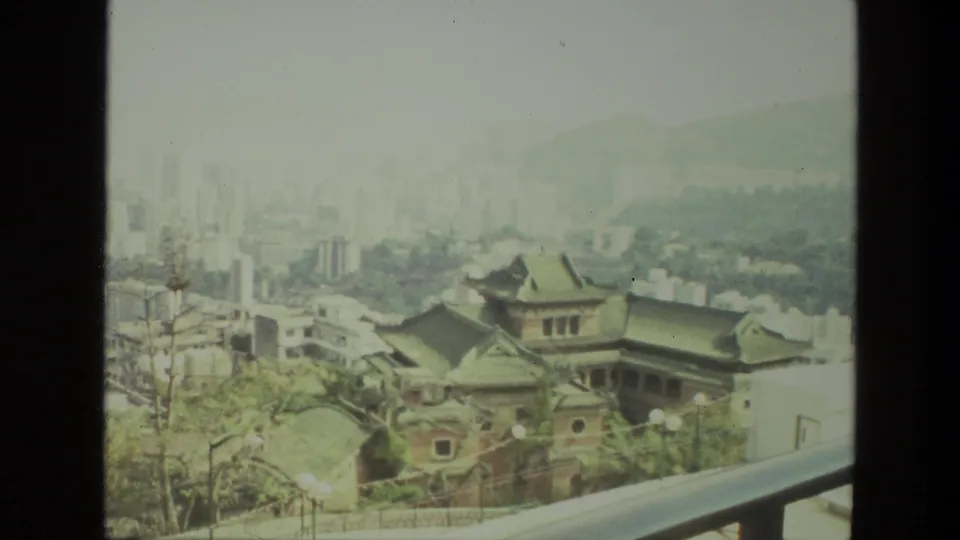 1974 India home-movie frame showing railing and traditional roofs in the foreground with a hazy city and hills beyond.
