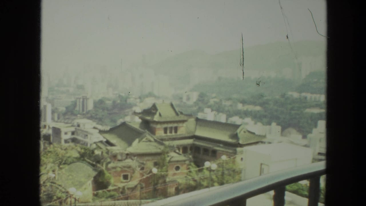 Hazy city view with tiled-roof buildings, white blocks, distant towers, green hills, and a balcony rail.