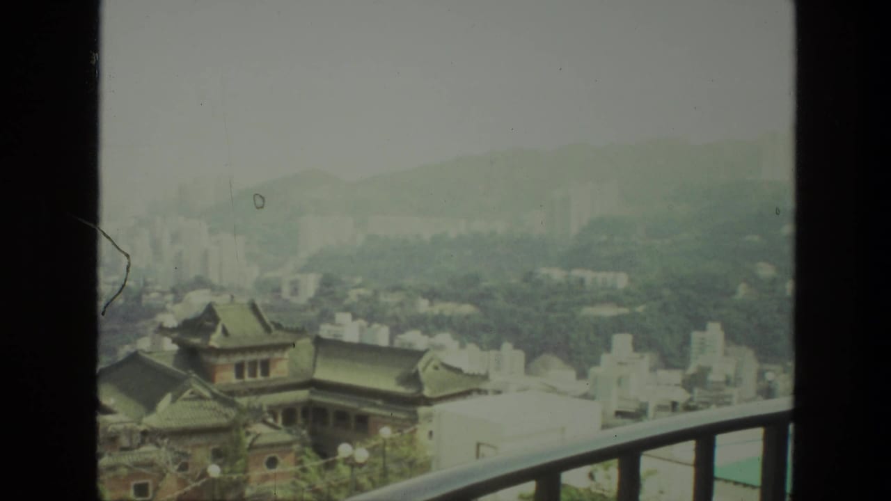 Hazy cityscape framed by a balcony rail, with tiled roofs, pale buildings, towers, and hills in the distance.