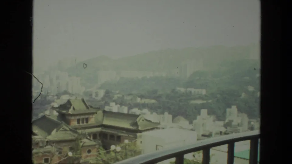 The 1974 8mm frame shows an elevated India city view with tiled rooftops, pale urban blocks, distant high-rises, and haze.