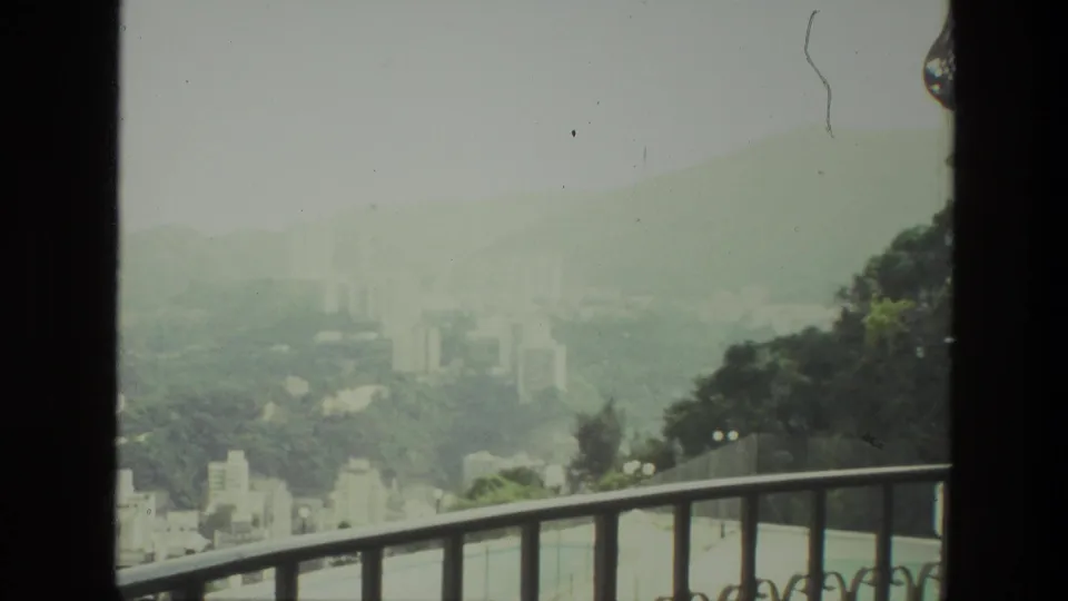 Hazy Indian cityscape in 1974, framed by a balcony rail and hillside greenery.