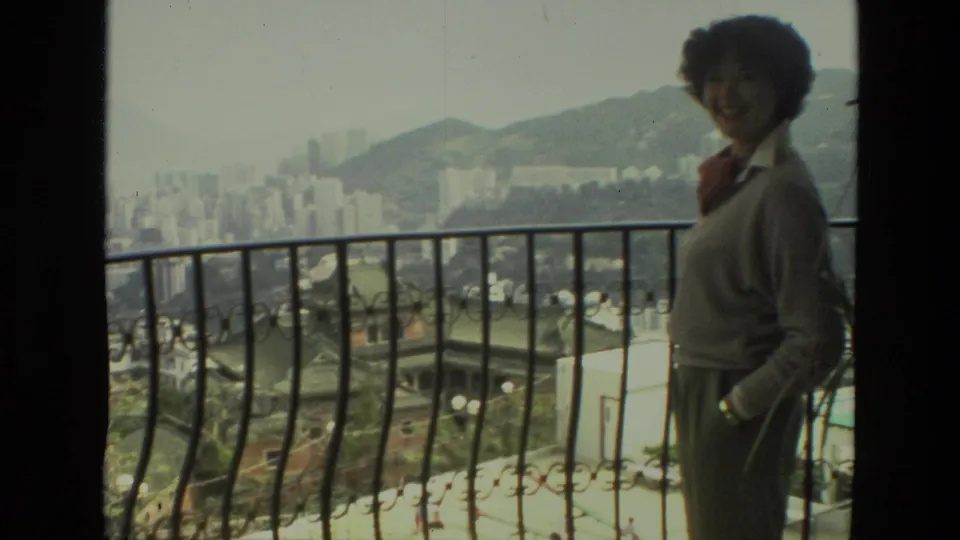 Adult woman in 1974 India home-movie footage stands by an ornate balcony railing above a hazy city and hills.
