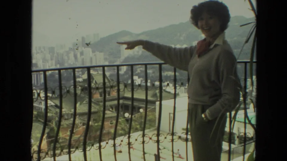 Adult woman in 1974 India home-movie footage smiles and points across the balcony toward the cityscape and hills.