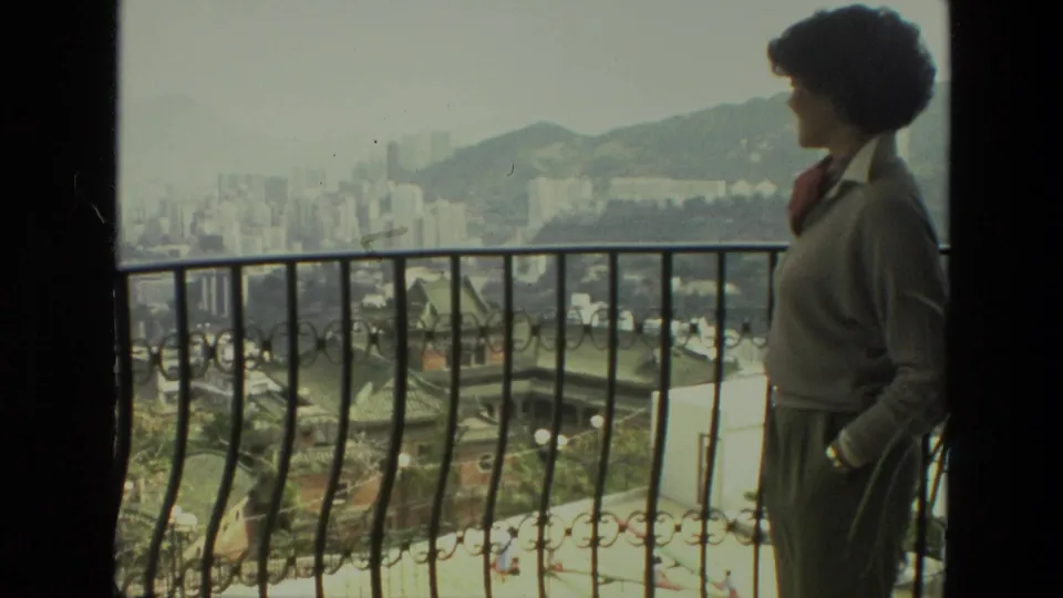 Adult woman in profile stands near an ornate railing in 1974 India footage, with a hazy urban hillside view behind her.