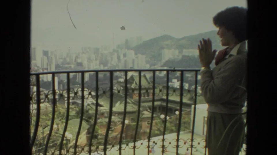 Adult woman in 1974 India footage stands partly at frame edge with hands clasped, overlooking a hazy city and hills.