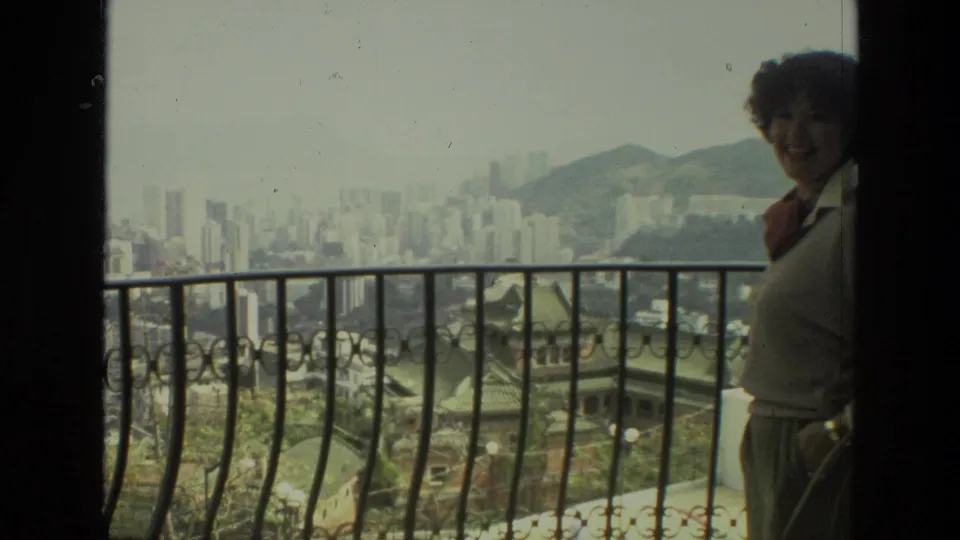 Adult woman in 1974 India footage smiles on a balcony above a dense cityscape with mountains in the distance.