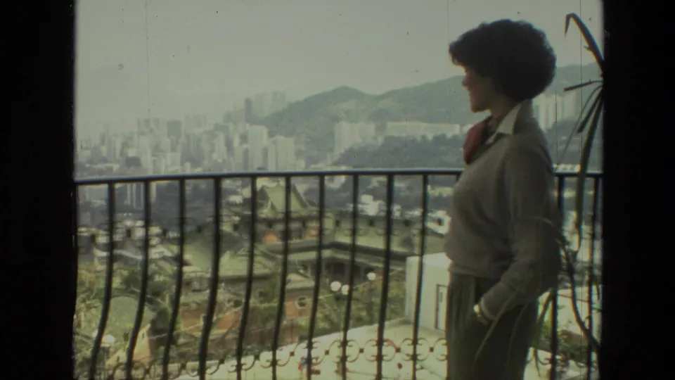 Adult woman in profile on a balcony, with 1970s India city buildings, rooftops, and hills visible beyond the railing.