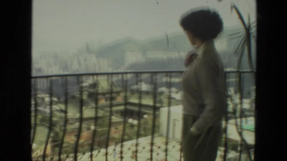 Adult woman moves along the balcony in soft 8mm focus, with a hazy city skyline and mountain backdrop visible in India footage.