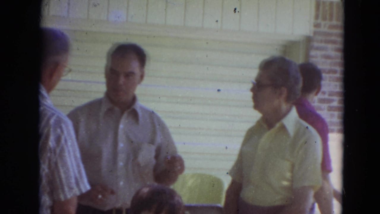 Men stand and talk outside a brick house near a garage door.