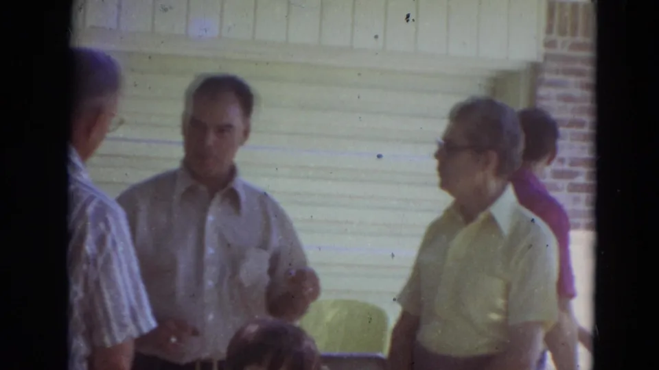 In 1974 Lincoln, Nebraska footage, several men stand outside a brick house near a garage door during an informal conversation.