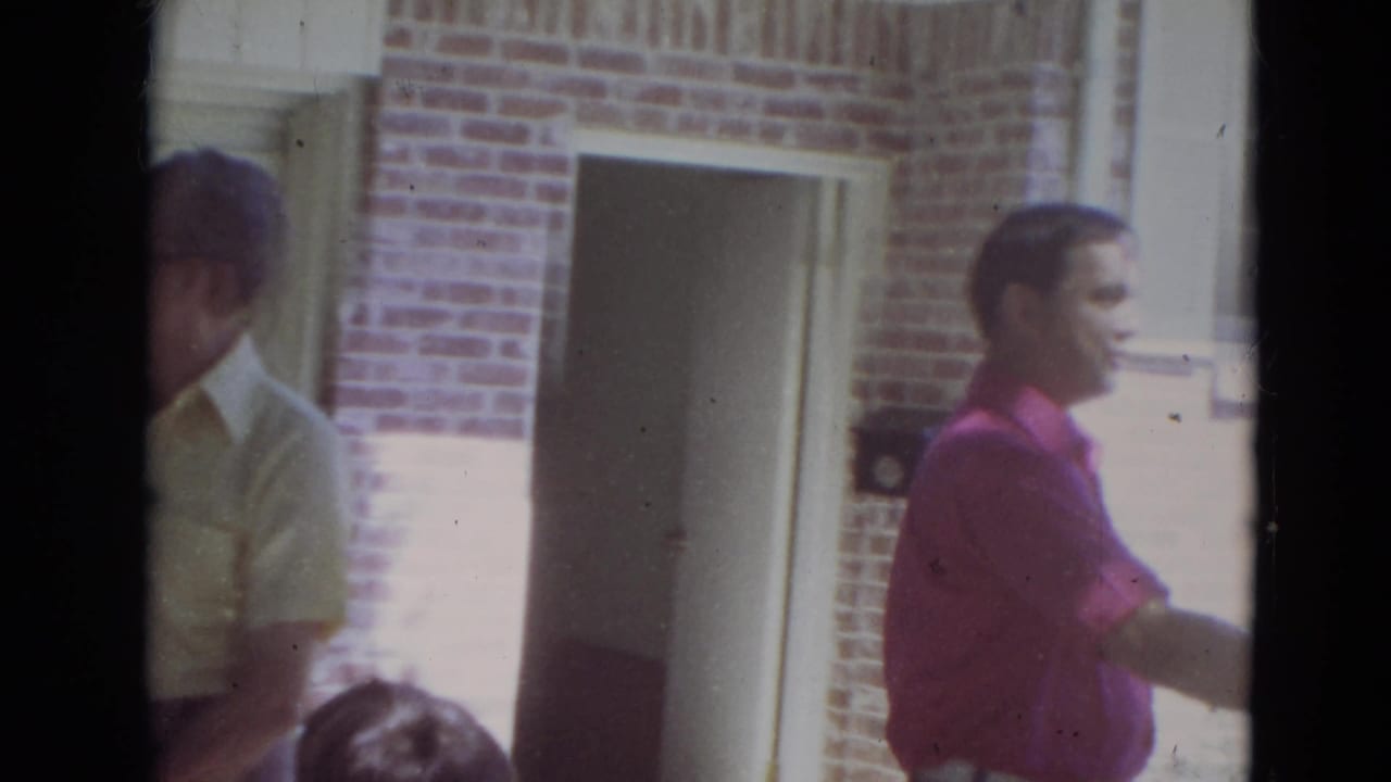 A man in a pink shirt walks past men outside a brick house.