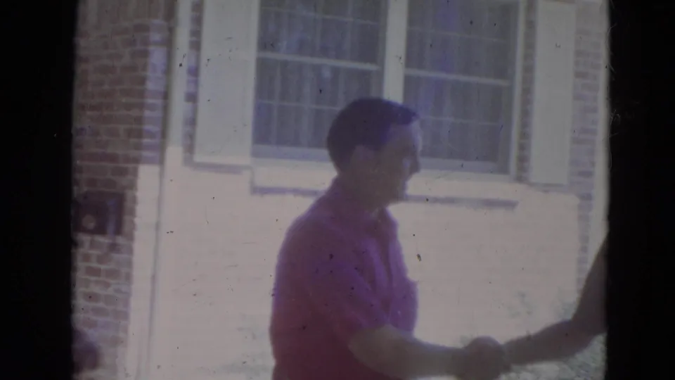 The man in a pink shirt begins a handshake outside the brick residential house in the 1974 Lincoln footage.