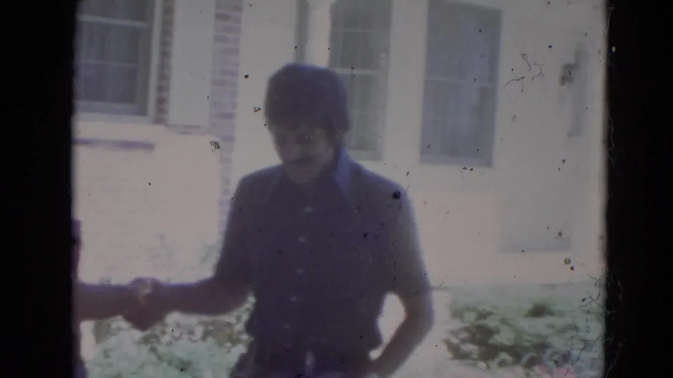 Two adult men shake hands outside a brick house, with windows and shrubbery behind them in the Lincoln, Nebraska home movie.