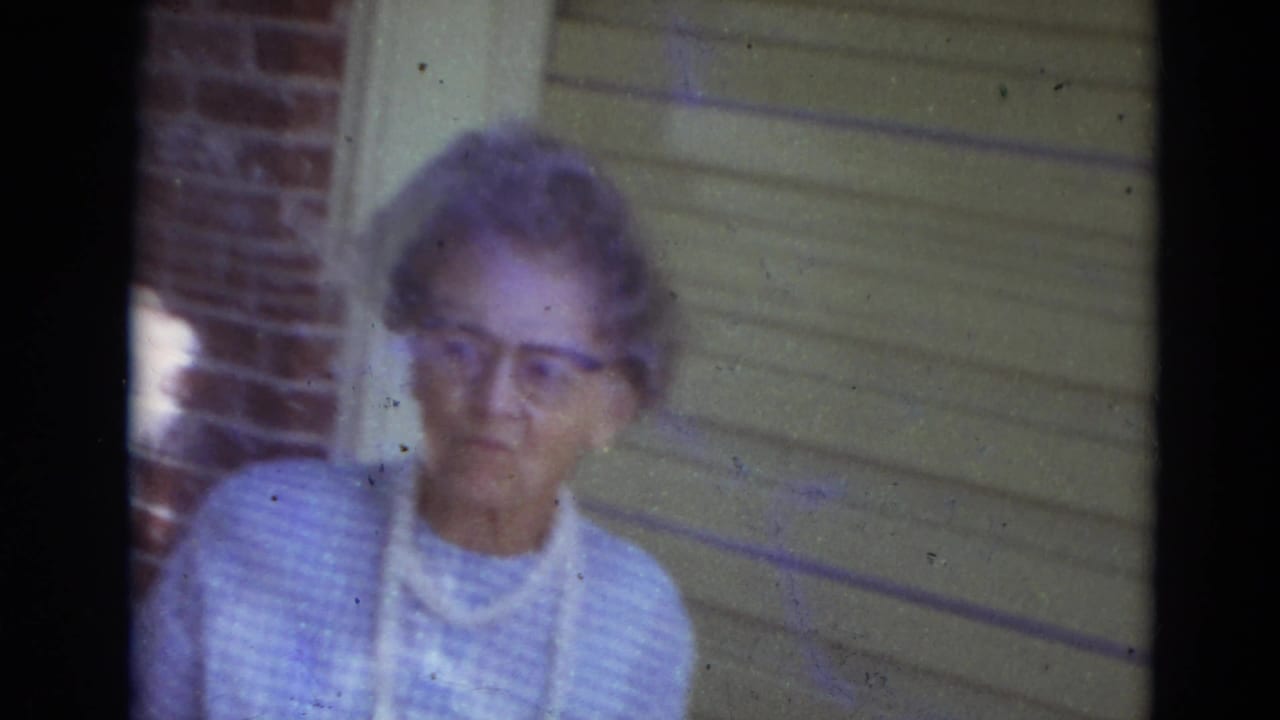 Elderly woman in a blue checked dress stands near brick and siding.
