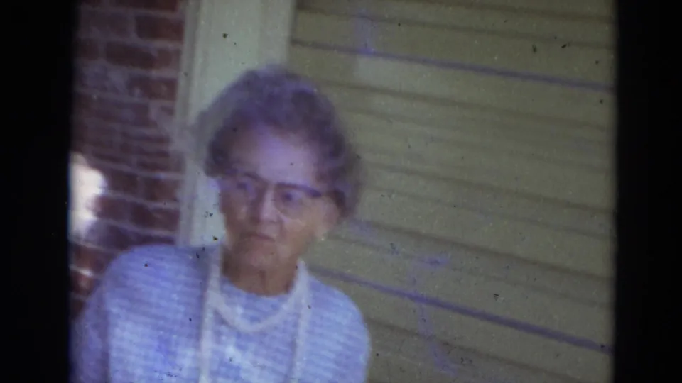 Elderly woman in Lincoln, Nebraska home-movie footage stands on a porch beside brickwork and light siding.