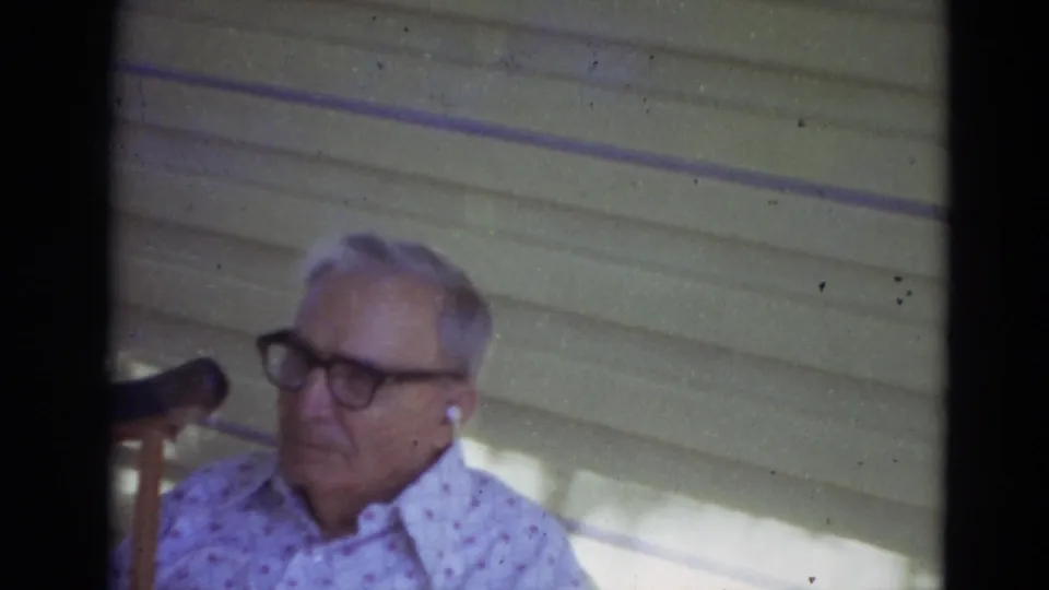 Older man with dark glasses and a patterned shirt stands near a porch chair in the 1974 Lincoln, Nebraska clip.