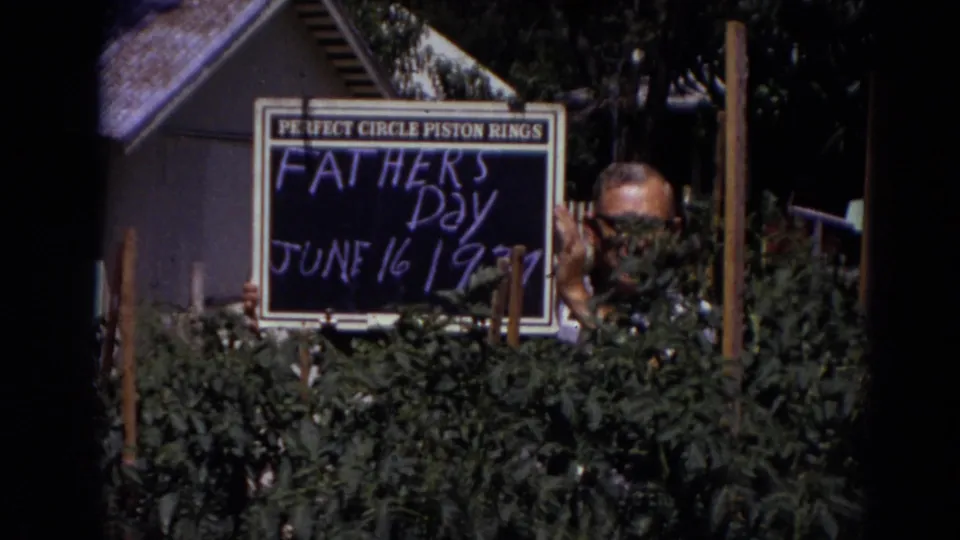 In Lincoln, Nebraska 1974 home-movie footage, a man in glasses stands behind shrubs with a dated Father's Day chalkboard.