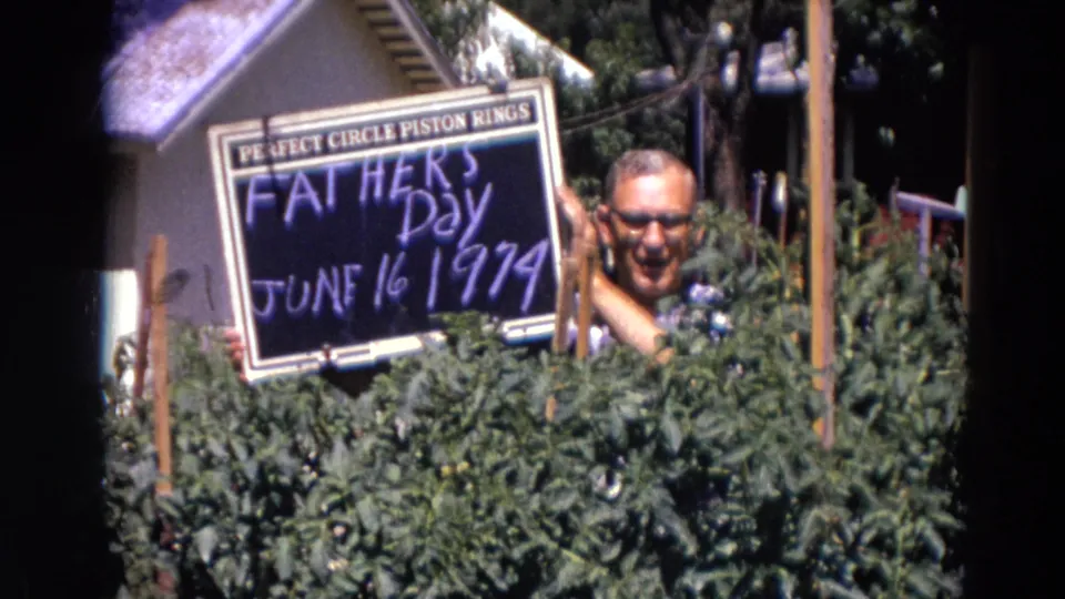 The smiling man raises a tilted Father's Day chalkboard above the shrubs in a 1974 Lincoln, Nebraska yard scene.