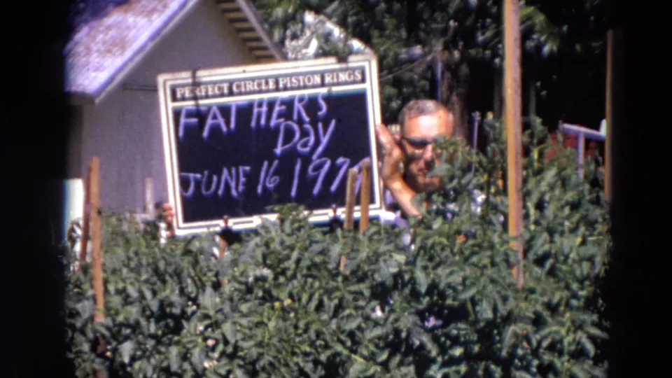 A man smiles beside the Father's Day chalkboard, with shrubs and house siding visible in 1974 Lincoln, Nebraska home footage.