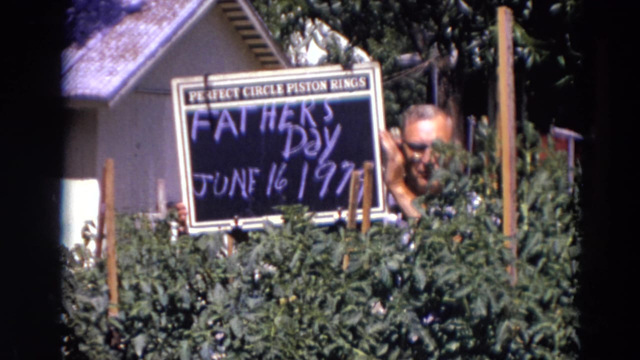 Man with glasses holds a Father's Day sign behind tall garden plants.