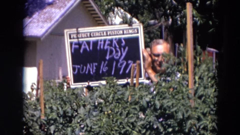 In Lincoln, Nebraska 1974 footage, a man with glasses smiles from behind garden plants while holding a Father's Day sign.