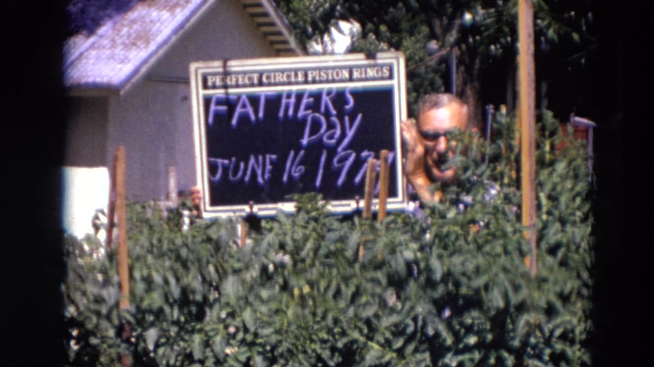 A smiling man appears beside a Father's Day sign in a sunlit Lincoln garden, with tomato plants and a house behind him.