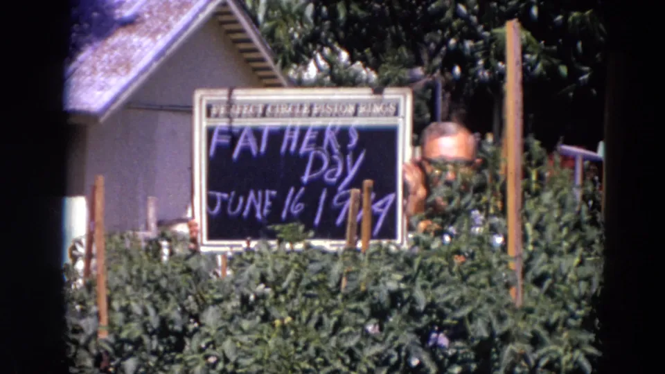 The man is partly obscured by tall plants as he holds the June 16, 1974 Father's Day sign near a house.