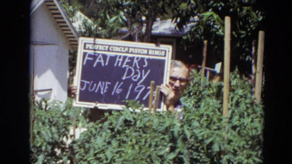 A man in glasses holds the Father's Day sign among dense garden plants in 1974 Lincoln home-movie footage.