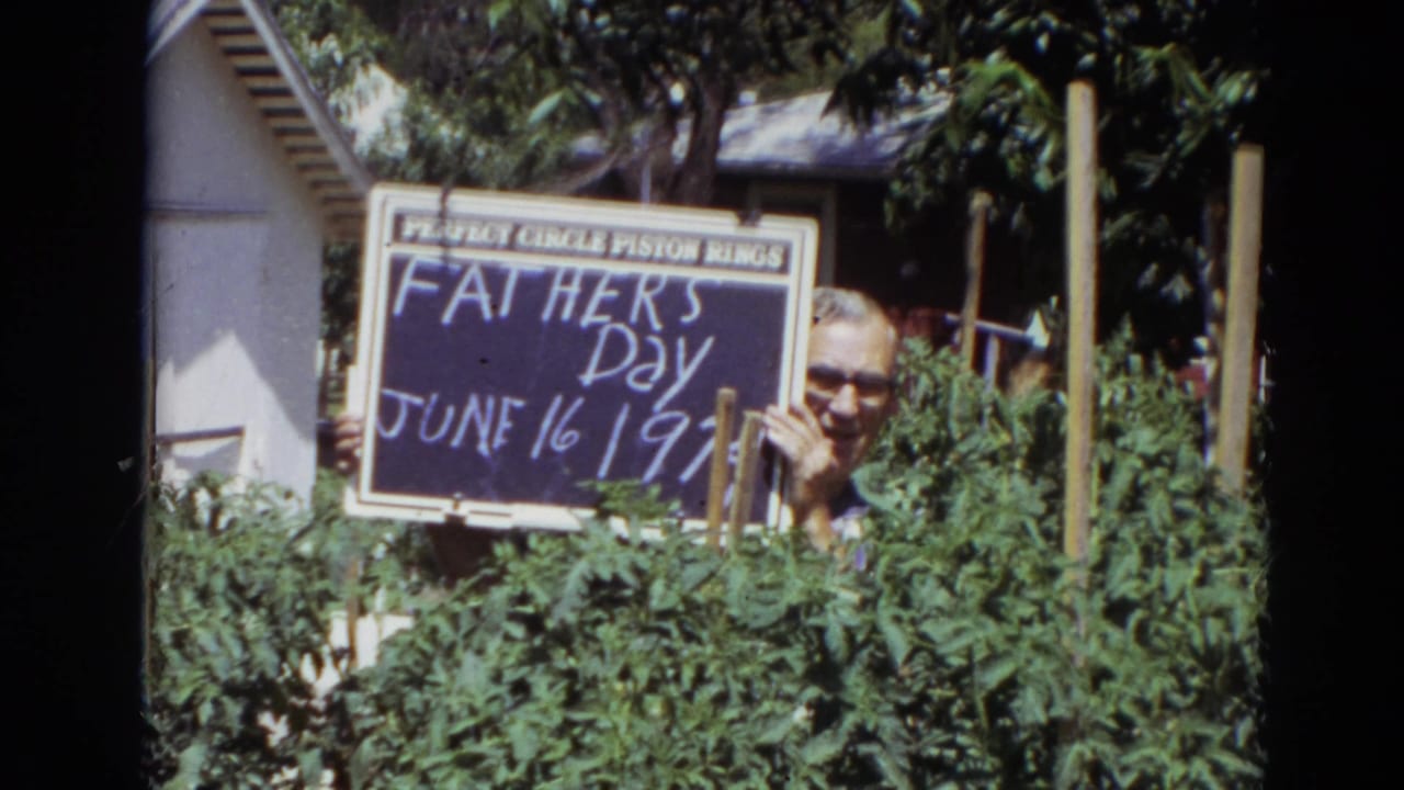 Man with glasses holds a Father's Day chalkboard sign among tall garden plants.