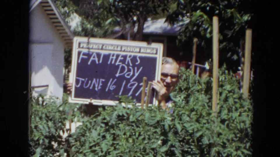 In 1974 Lincoln, Nebraska home-movie footage, a man with glasses holds a Father's Day chalkboard sign while partly hidden by tomato plants.