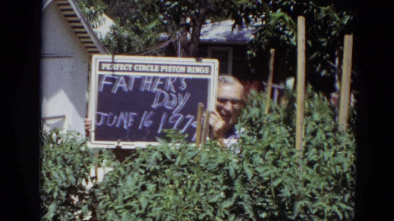 Man behind garden plants holds a Father's Day sign with piston rings text at top.